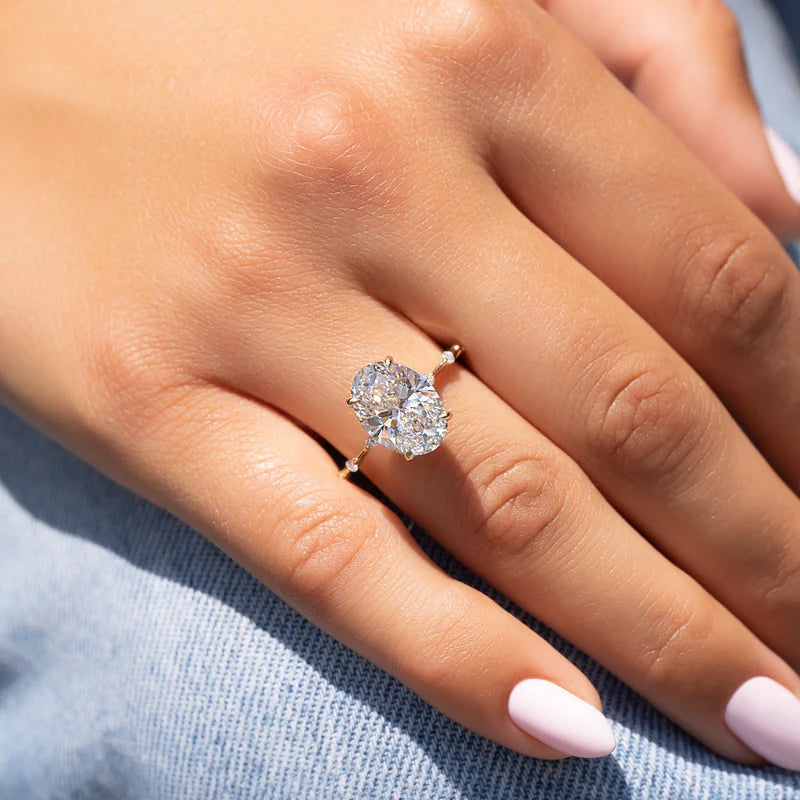 A close-up of a woman’s hand wearing a two-stone oval diamond engagement ring set in yellow gold, featuring brilliant-cut ovals arranged side by side on a delicate band, styled with soft pink nails and natural daylight.