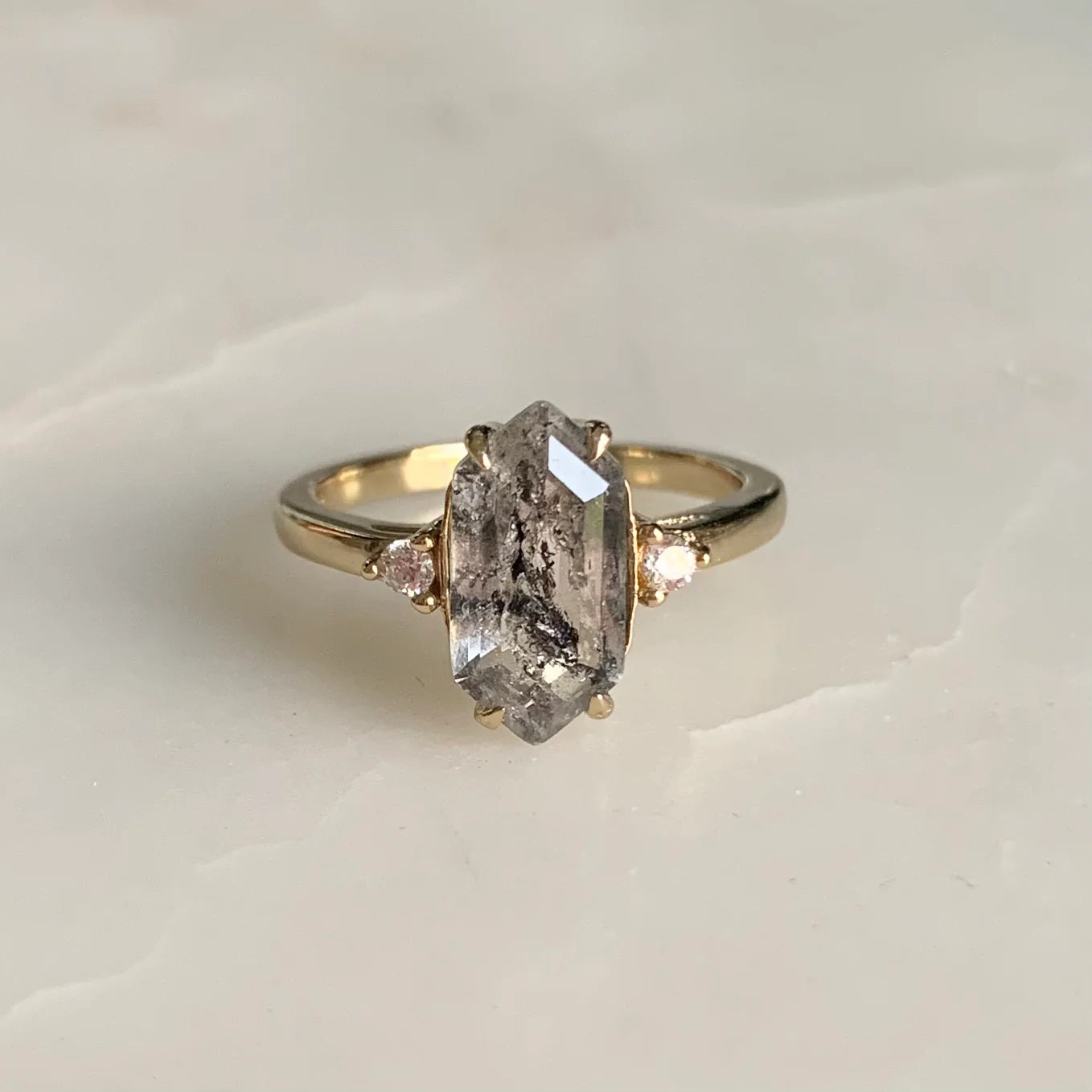 Salt and pepper diamond engagement ring featuring a long hexagon-cut stone with bold black inclusions, set in a yellow gold band with side diamonds, styled against a soft beige surface.