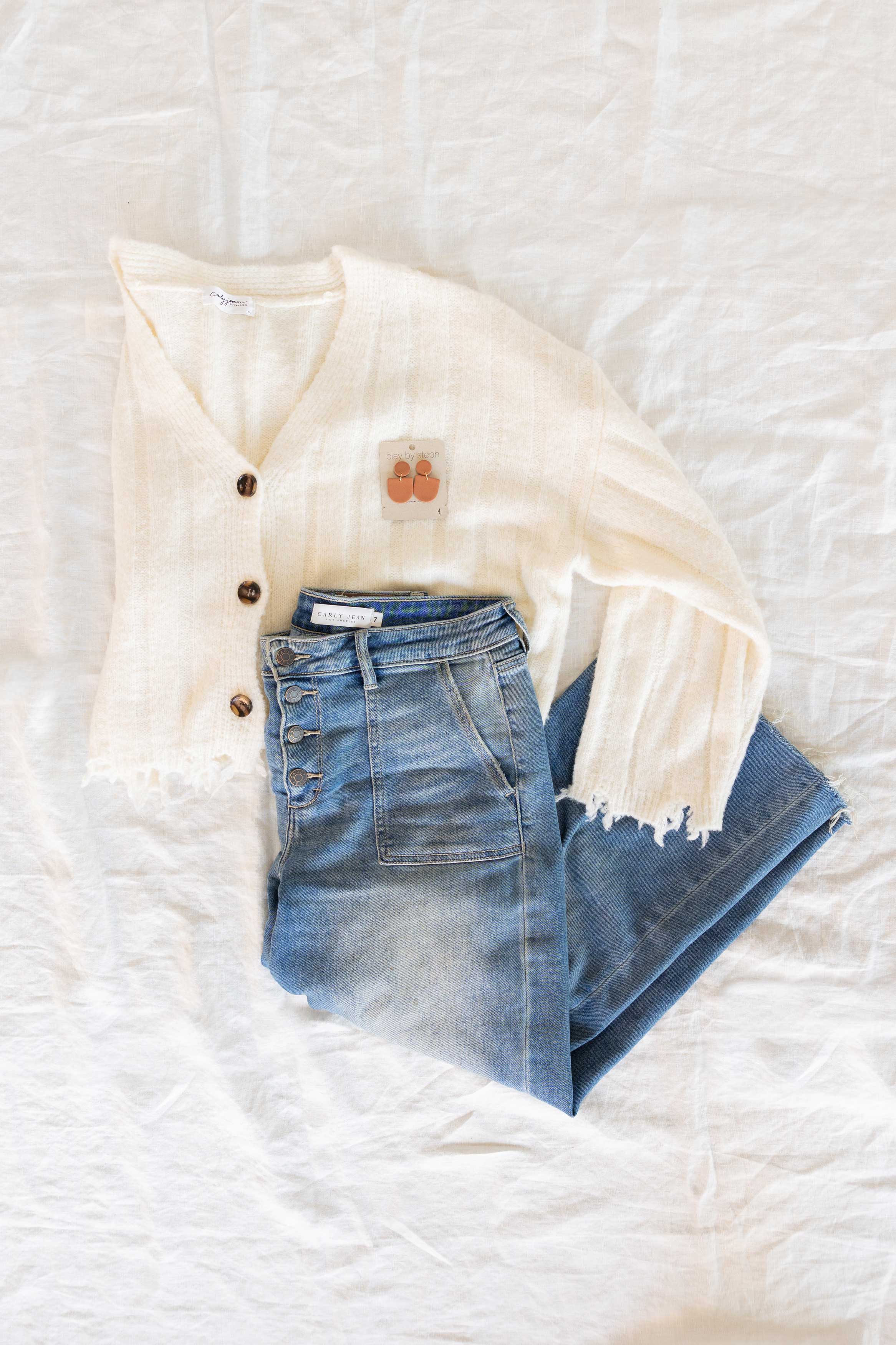A folded white cardigan and folded blue jeans lay on a bed with Pumpkin Spice Mini Camille clay earrings.
