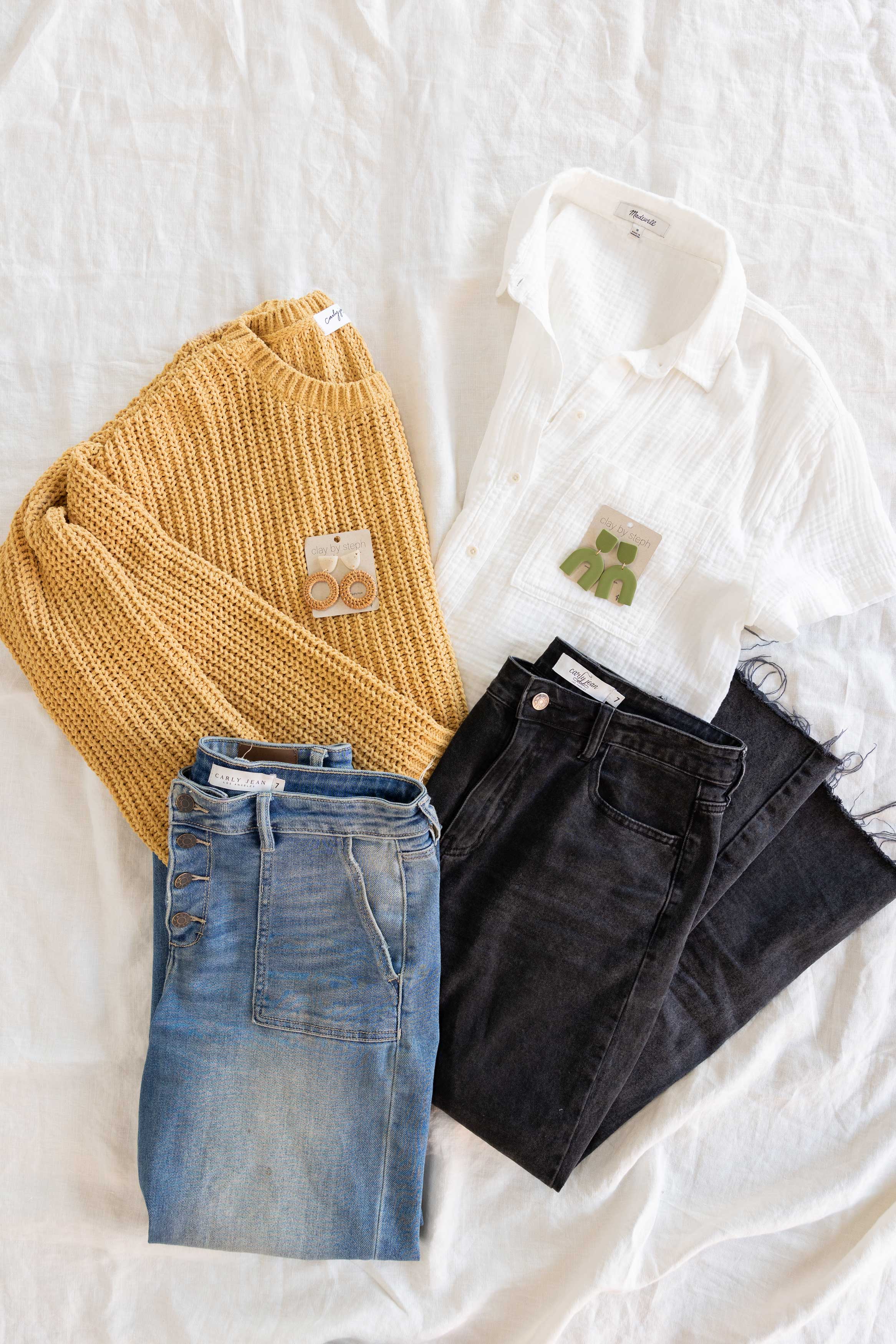 Two folded outfits lay on a bed. One is a folded marigold sweater, blue jeans, and Sea Salt Britta clay earrings. The second is a folded white linen shirt with black jeans and green clay earrings.