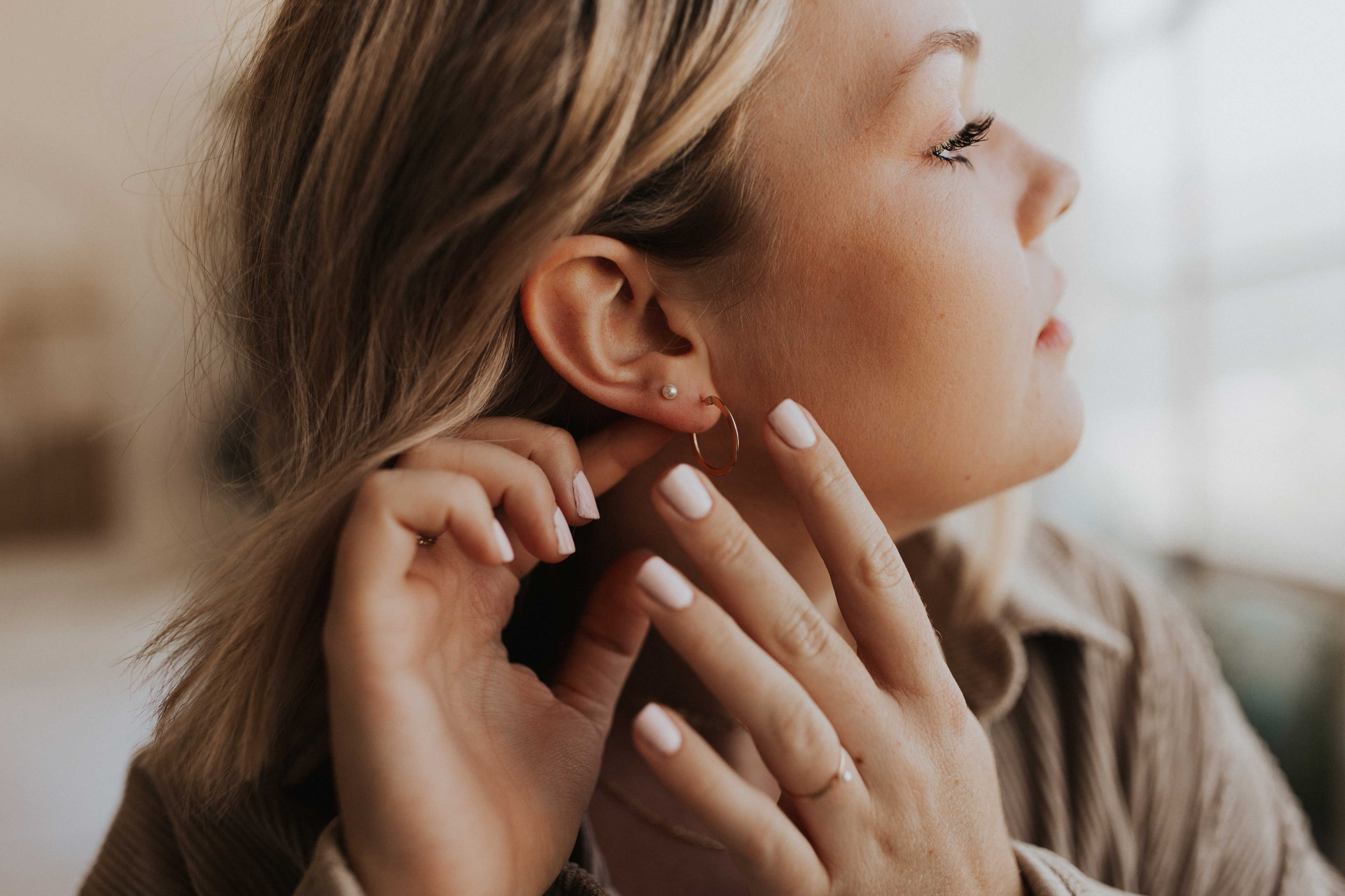 A blonde woman is putting on a gold hoop earring while wearing a smaller pearl stud in a second piercing. 