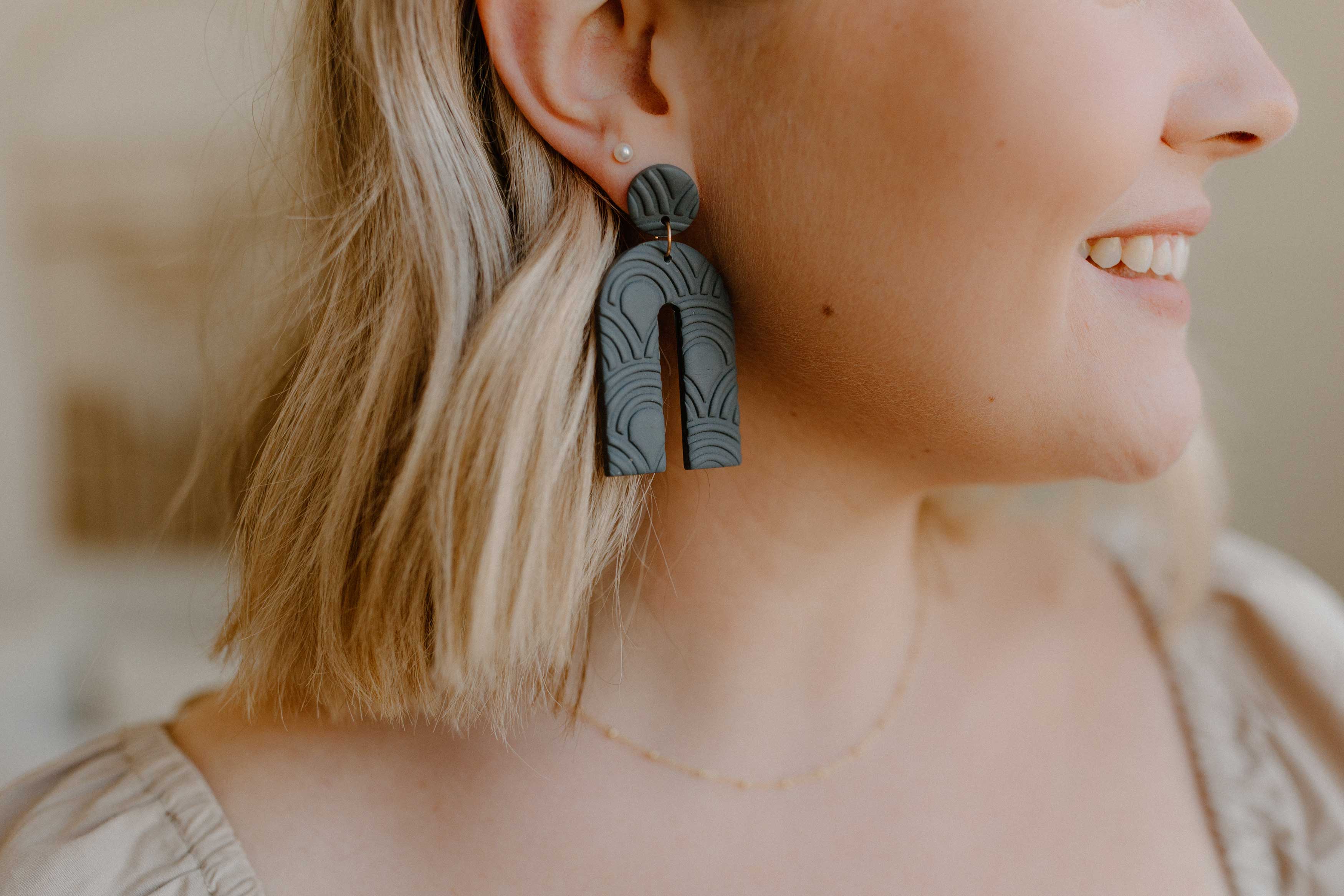 A blonde woman smiles showing off arch-shaped Charcoal Wren clay earrings.