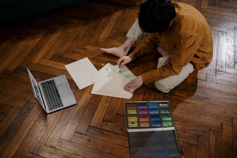 A Person Sitting on the Wooden Floor While Making a Drawing with Crayons on the White Paper
