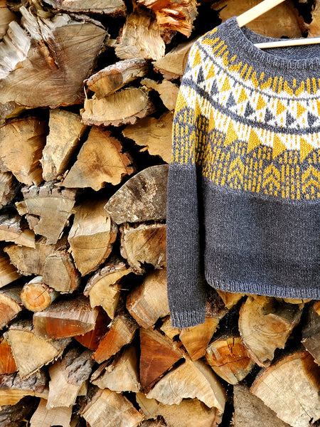 a hand knit colorwork sweater pattern made from American organic cotton + merino wool posed against a wall of firewood