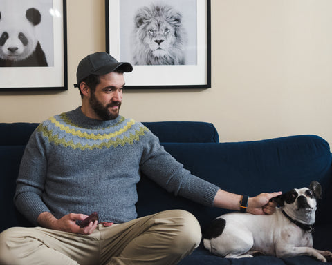 A bearded white man wears a handknit colorwork sweater on a couch petting a super cute black and white dog 