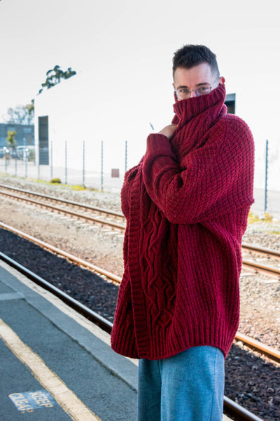 A white man with short dark hair wears a red handknit wool cardigan with cables as he playfully hides his face in the high collar