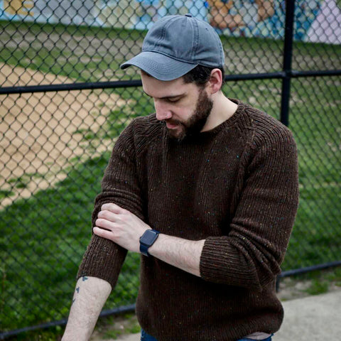 A bearded white man in a blue baseball cap pulls up the sleeve on his handknit brown sweater and regards the fabric