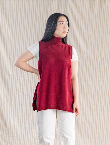 Rebekah, a woman with long straight black hair, wears a red turtleneck tunic vest and looks away from the camera to the right.