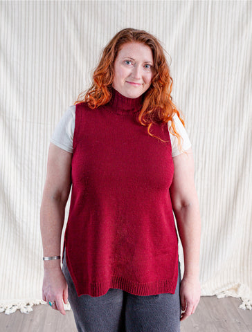 Tammy, a woman with long red-brown curly hair, wears a red turtleneck tunic vest and looks into the camera.