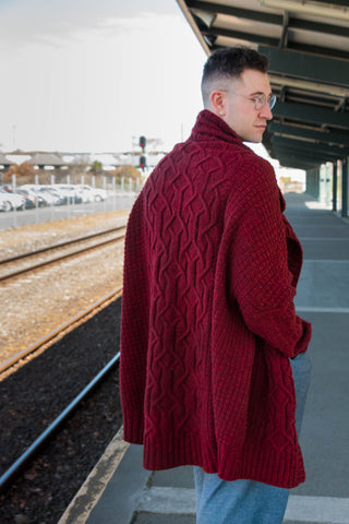 A long red cabled handknit cardigan is worn by a white man with his back turned to the camera, looking over his shoulder