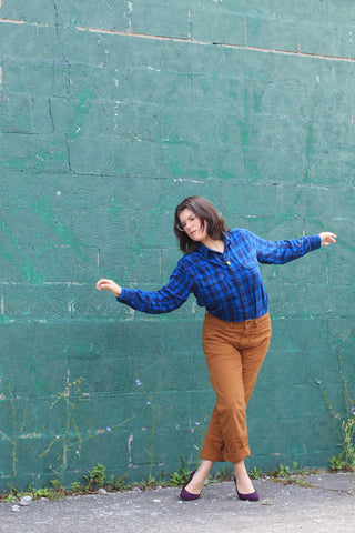 A white non-binary person in a blue flannel arcs their body in a dancerly pose in front of a teal brick wall.