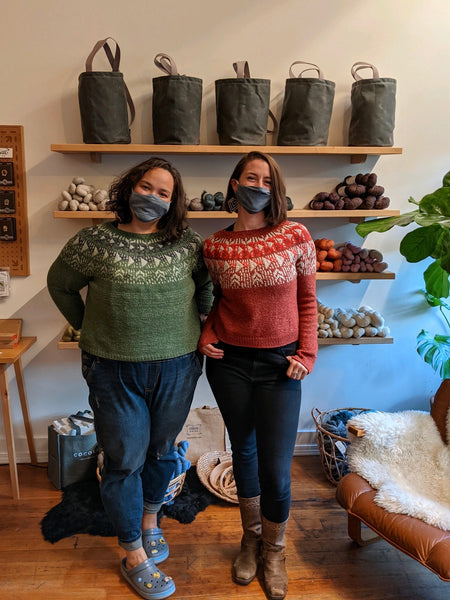 Two women pose in a yarn store wearing matching hand knit colorwork sweaters in two different colors. 