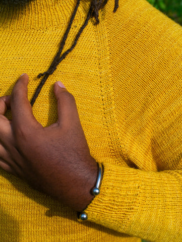 A close cropped image of a Black man with locs wearing a bright yellow handknit raglan sweater.