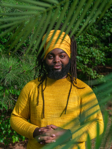 A Black male knitter, crocheter and educator wears a handknit happy yellow raglan sweater, tucked back behind tropical palm leaves.