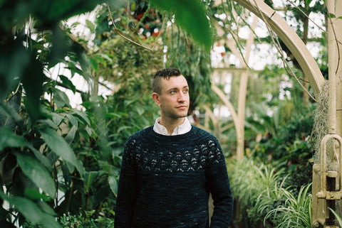 A dark teal handknit sweater with a lace yoke is worn by a white man with dark hair against a background of large plants