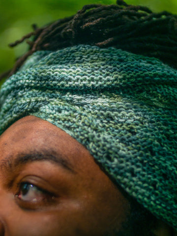 A teal handknit scarf is knotted at the top of a Black man's brow with locs peeking out the top.