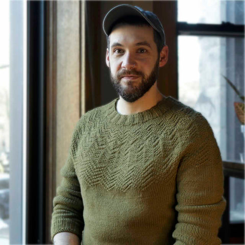 A white man with dark beard wears a handknit green sweater and looks into the camera with a friendly expression