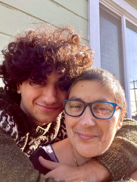 Emanuela sits in the sunlight outside her home. Her son stands behind her hugging her.