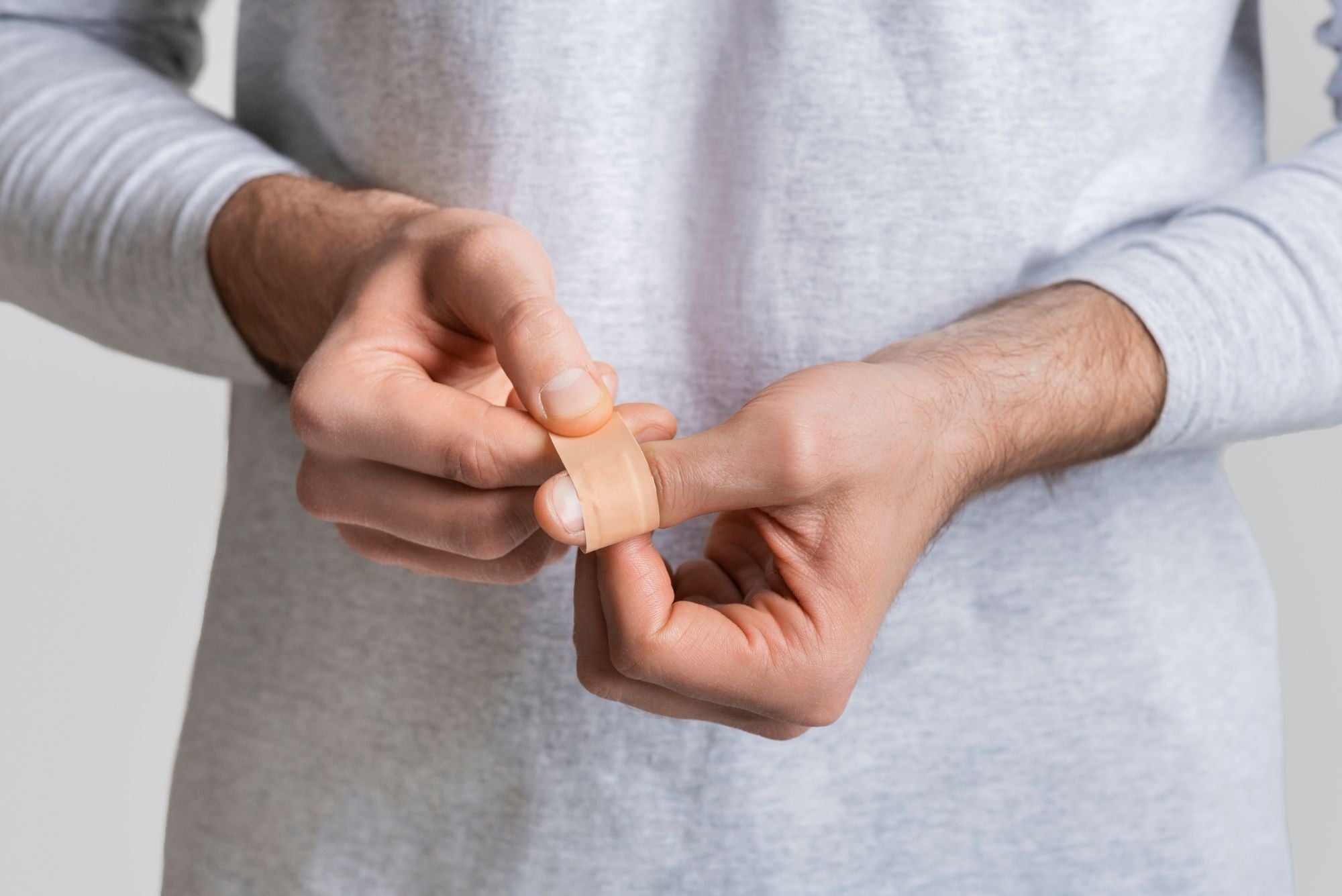 A person in a grey shirt stands in front of a white wall wrapping a band-aid around herpetic whitlow on their left thumb.