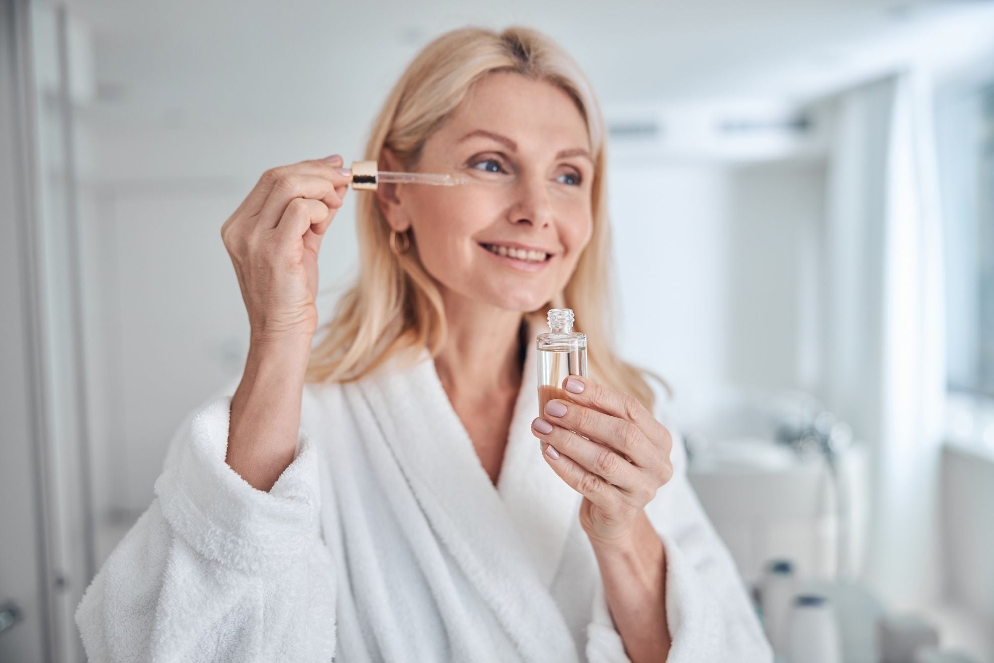 A blonde woman wearing a white robe prepares to use a dropper of essential oil on her canker sore.