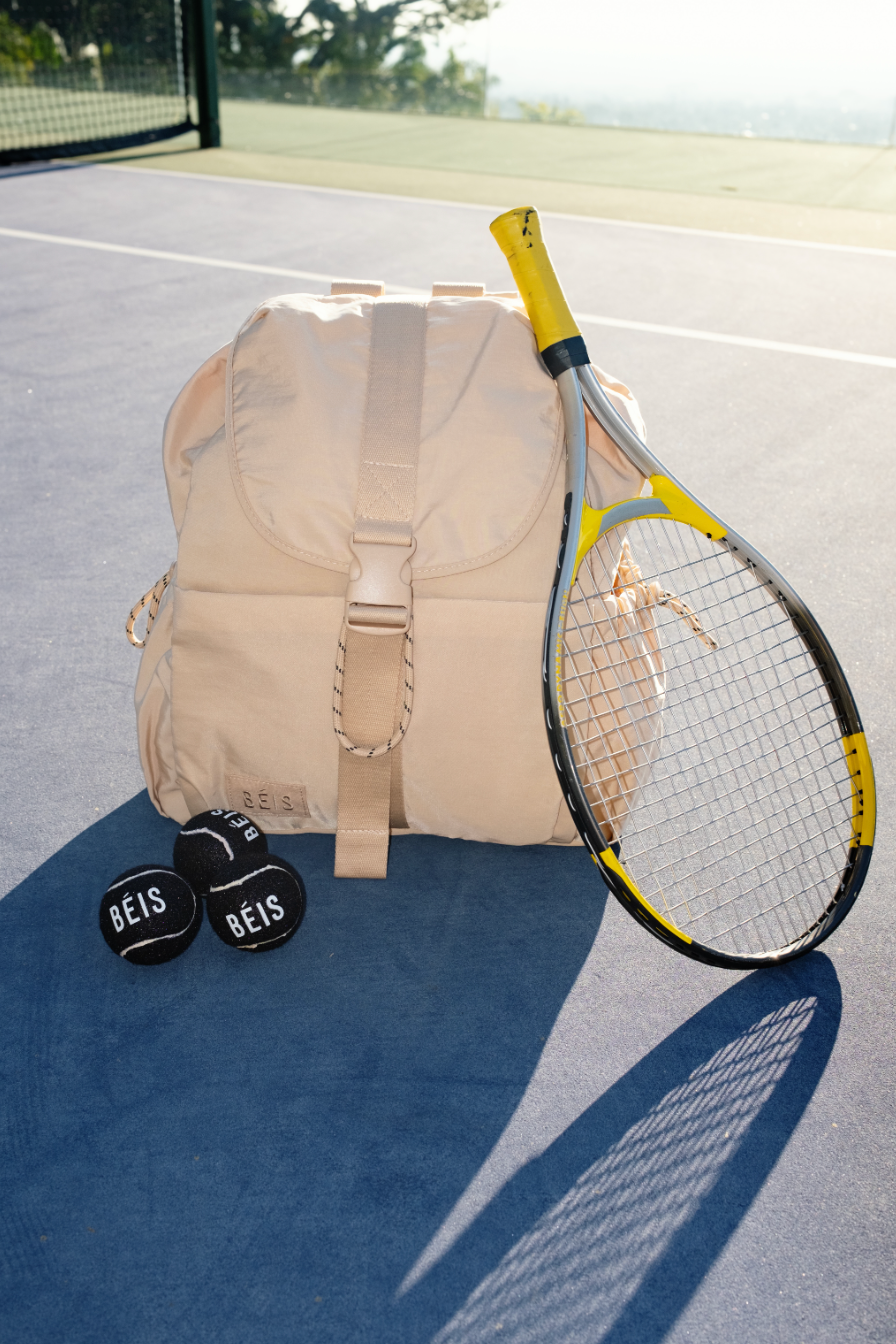 beige sport backpack sitting on a tennis court with a tennis racquet leaning on it