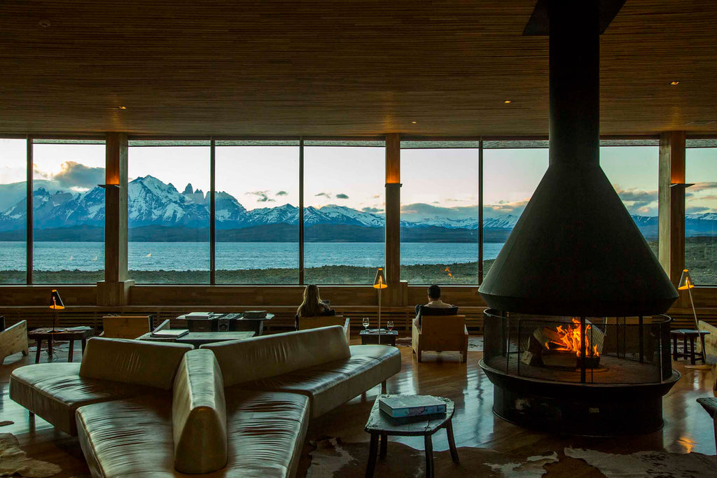 A view from inside the lodge of Tierra Patagonia in Chile. An open fire is roaring and a couple are looking out to mountain views 