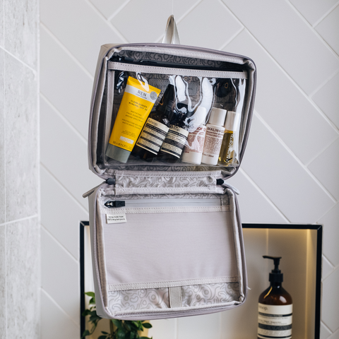 grey OneNine5 wash bag zipped open and hanging up in a bathroom. Toiletries and cosmetics are packed inside