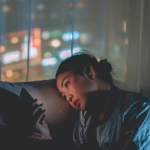 a woman stares at her phone with blue light in a dark room as the city lights blur outside the window