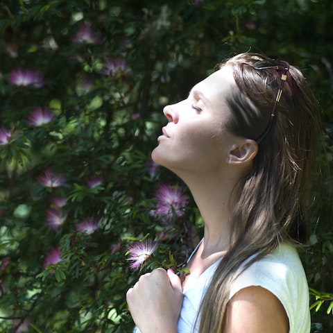 A woman with her eyes closed is enjoying sun on her face against a backdrop of green leaves and purple flowers