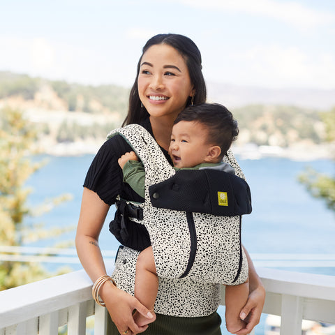 Mother with baby in a Lillebaby carrier. She's standing on a balcony with a view of a lake and forest behind her.