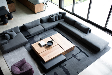 A birds eye view of a luxury living room with a handmade gray rug with a large rug sectional couch sitting on top. There is a large wooden coffee table in the center with magazines and a black bowl on top. 