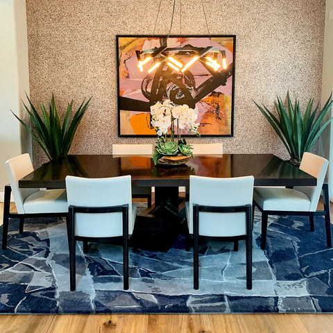 View of a modern dining room with a black table and white chairs on top of a handmade rug. The rug is blue with a light gray and blue tape like abstract design and a pink abstract painting on the wall.