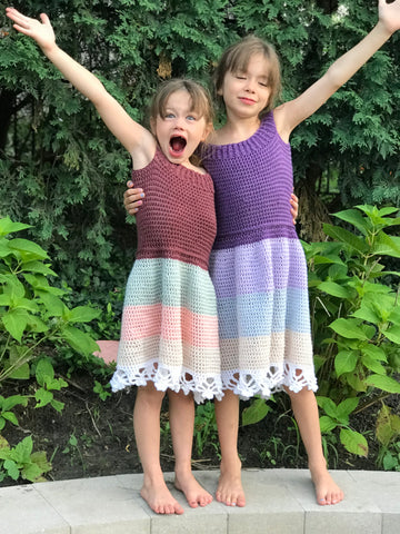 two young girls wearing crochet dresses with one arm around each other & the other up in excitement