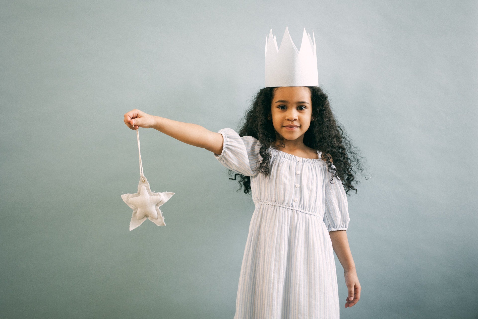 A cute little girl in a white costume with a paper crown on her head holding a white star on a string.