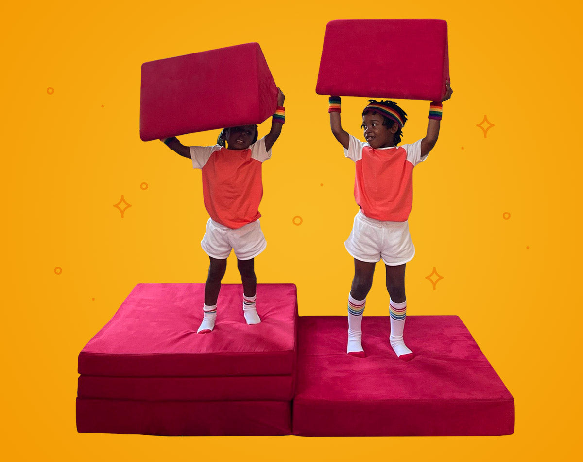 Two children standing on Nugget cushions, holding pillows overhead like weightlifters