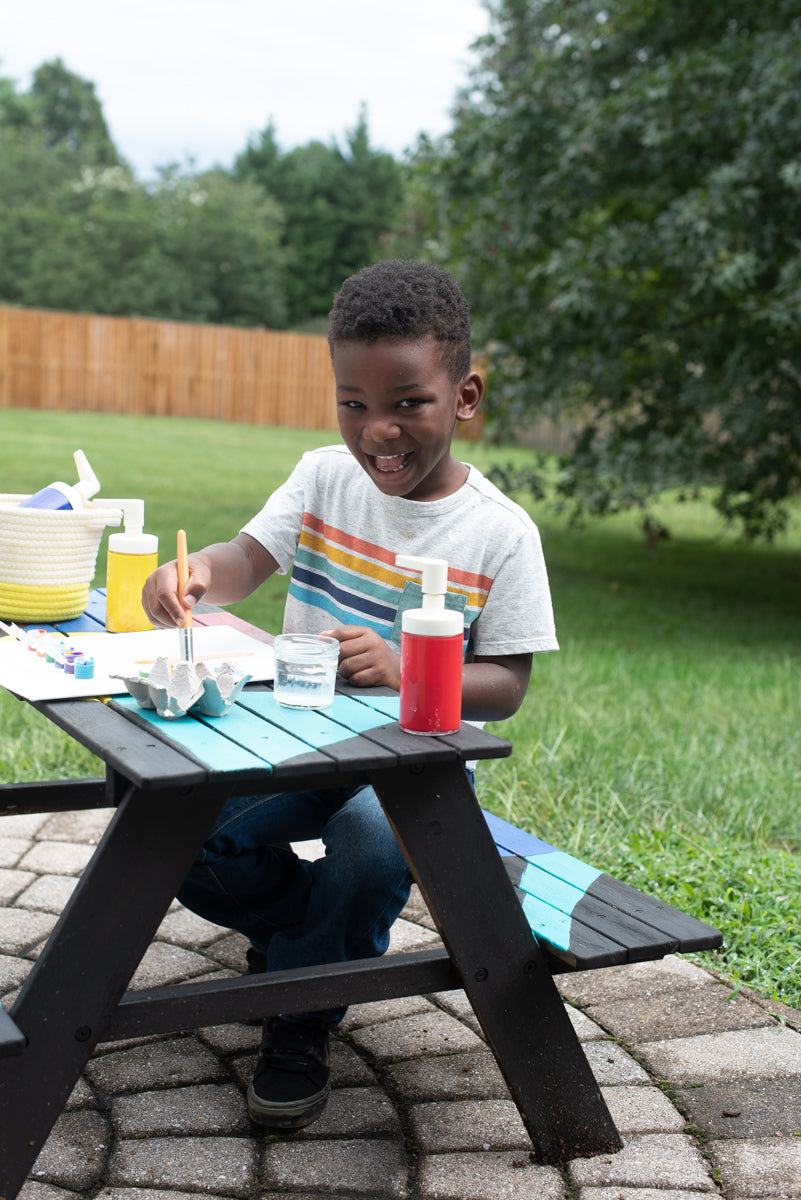 Jonah seated at an outdoor picnic table, painting and grinning at camera.