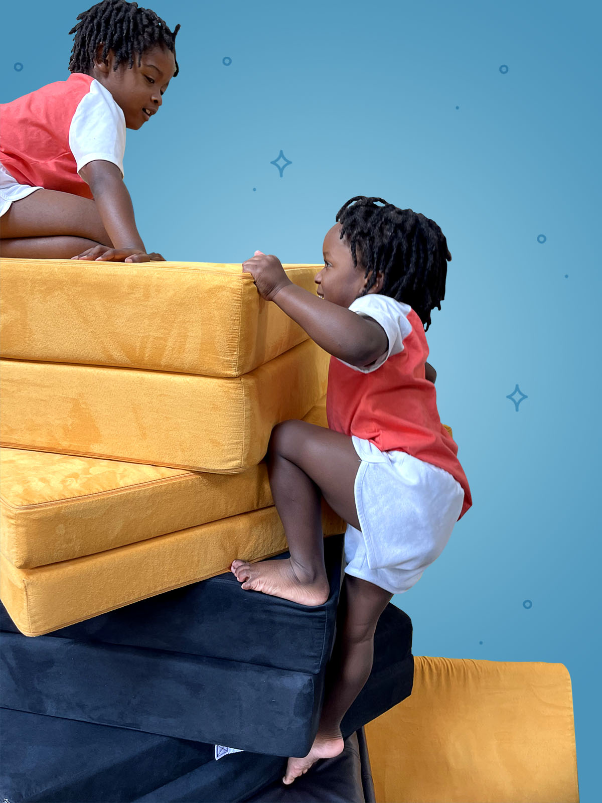 One child perched on top of a stack of cushions, looking down at other child who is climbing up the stack like a rock climber