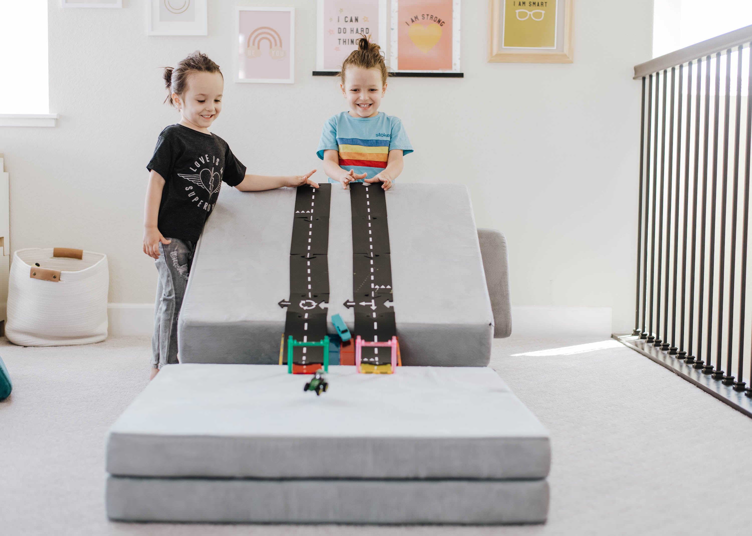 Two children standing behind a Nugget car ramp, with toy tracks laid down on one Nugget piece and cars race down towards another Nugget piece.