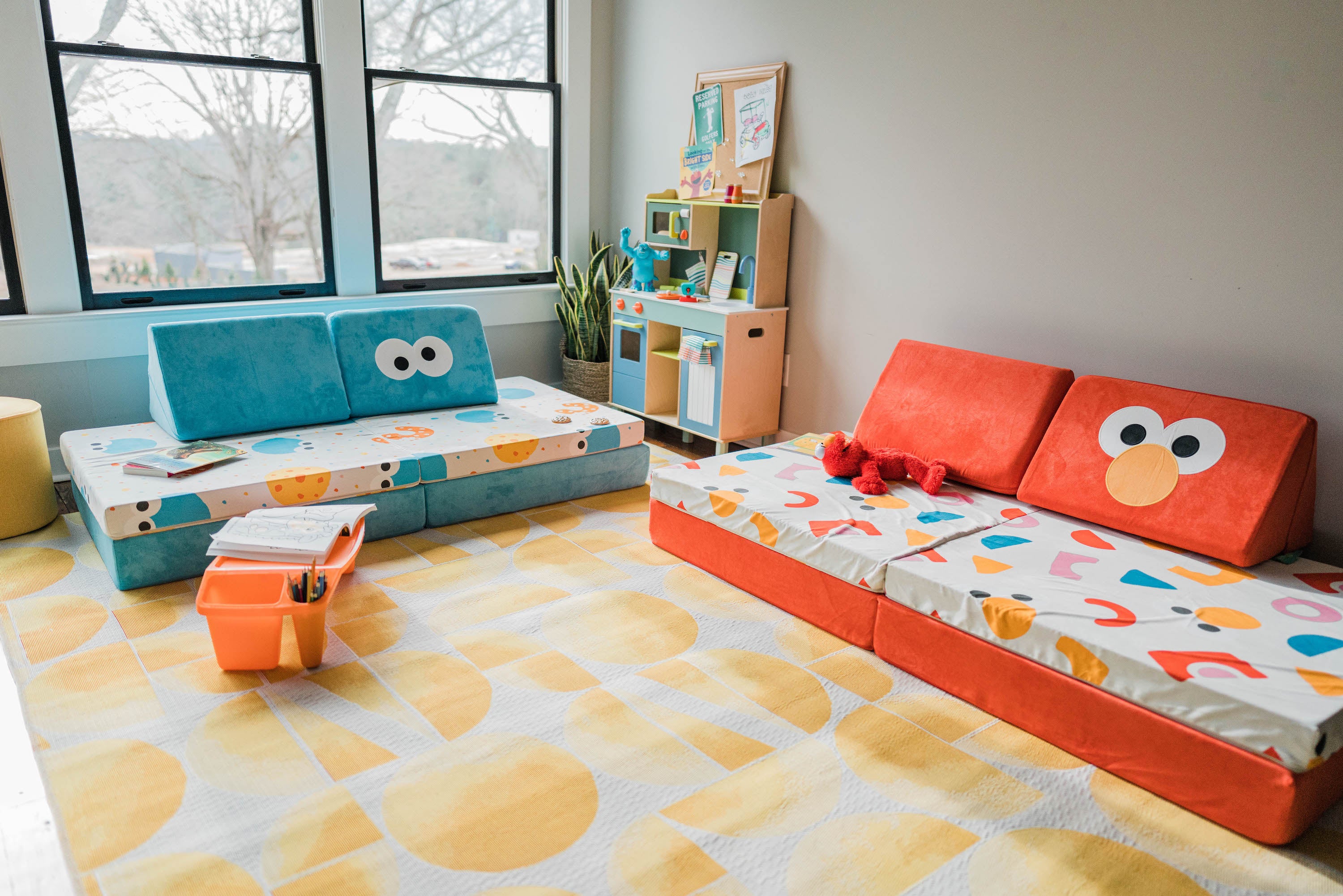 The Cookie Monster Nugget and Elmo Nugget in a playroom with a cream and yellow rug, a play kitchen, and assorted toys.