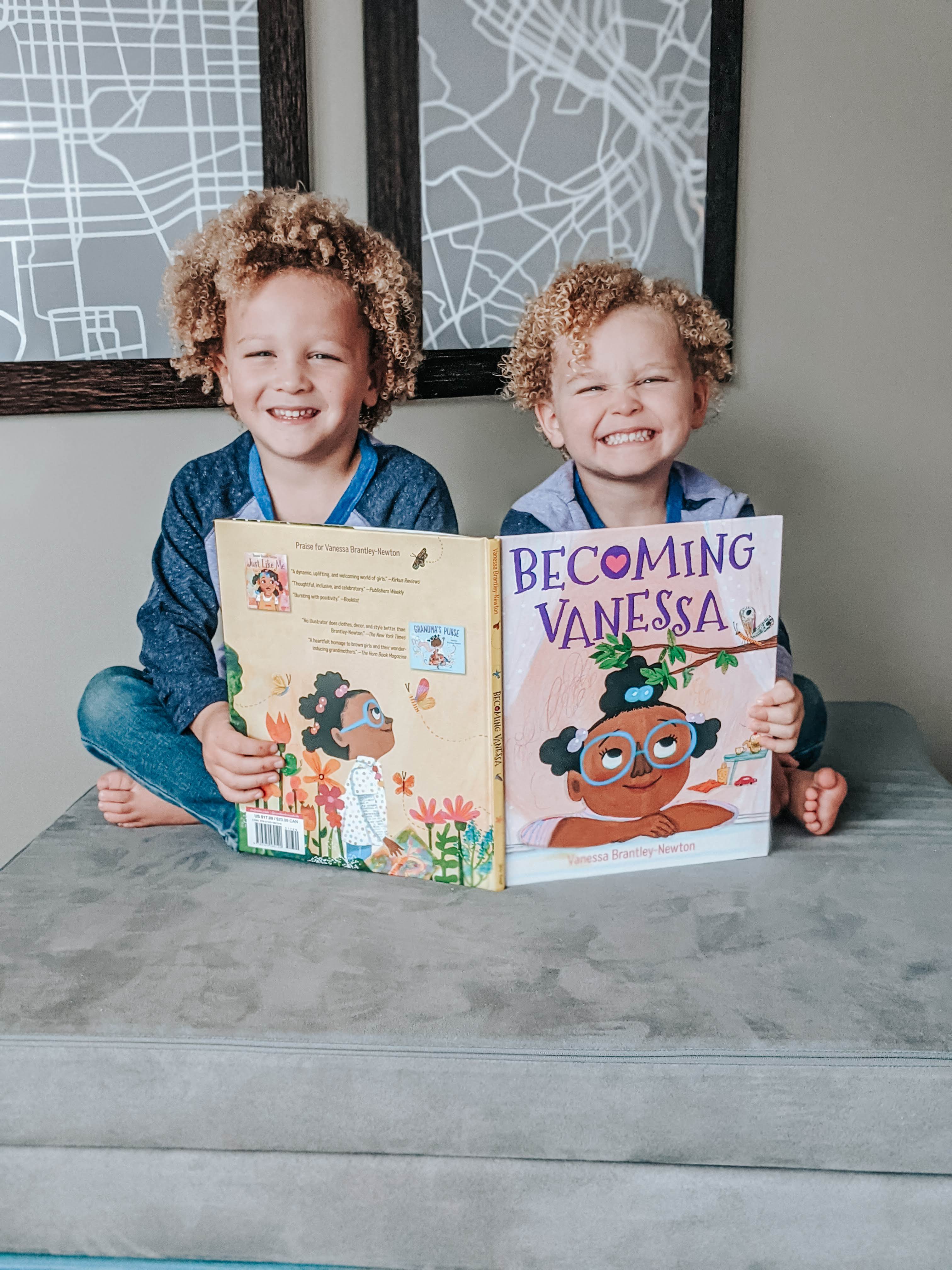 Two curly-headed kids holding "Becoming Vanessa" book open while sitting on a Nugget, grinning at camera