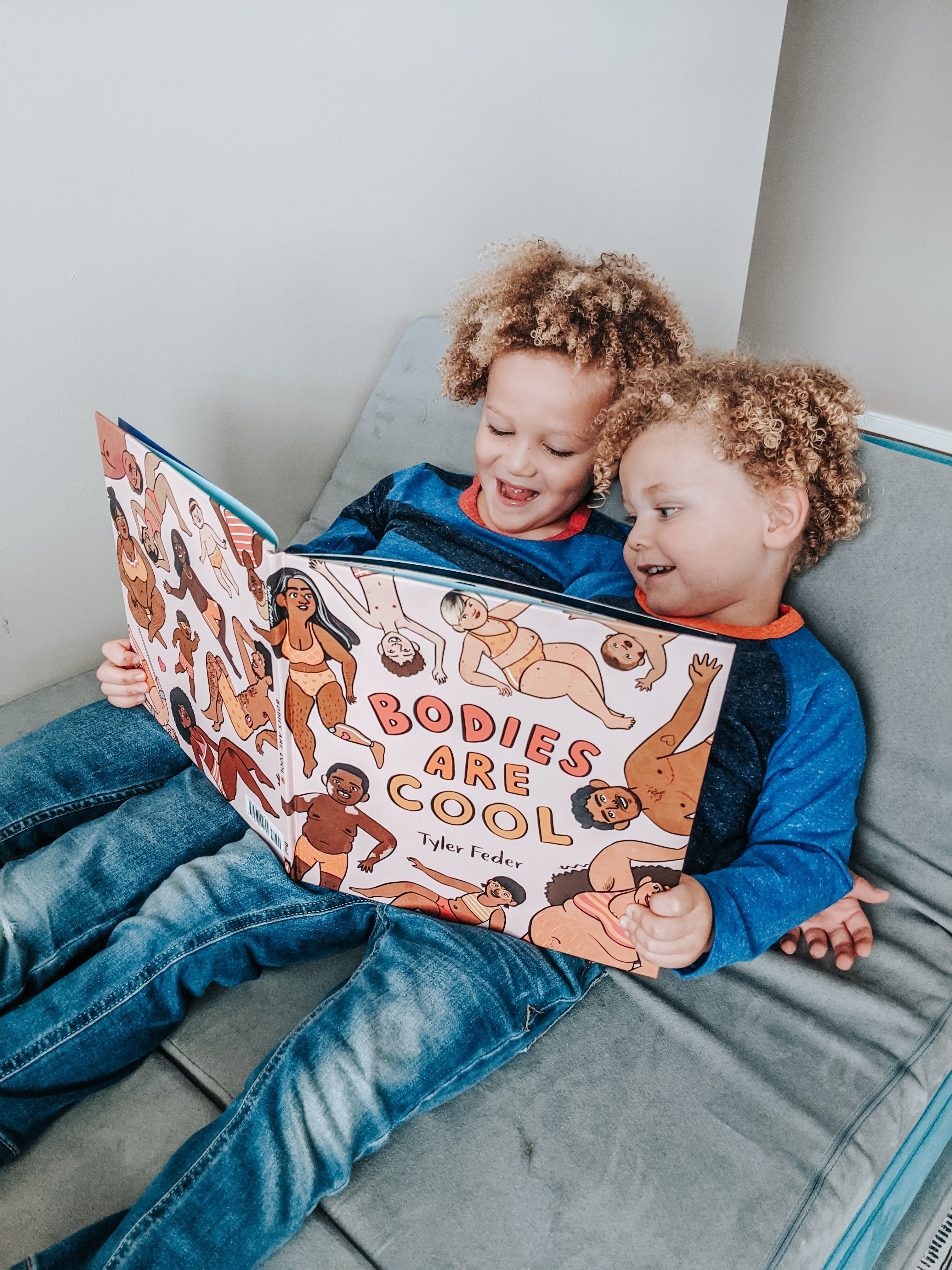Two kids leaning back on a Nugget couch, holding and reading "Bodies are Cool" together