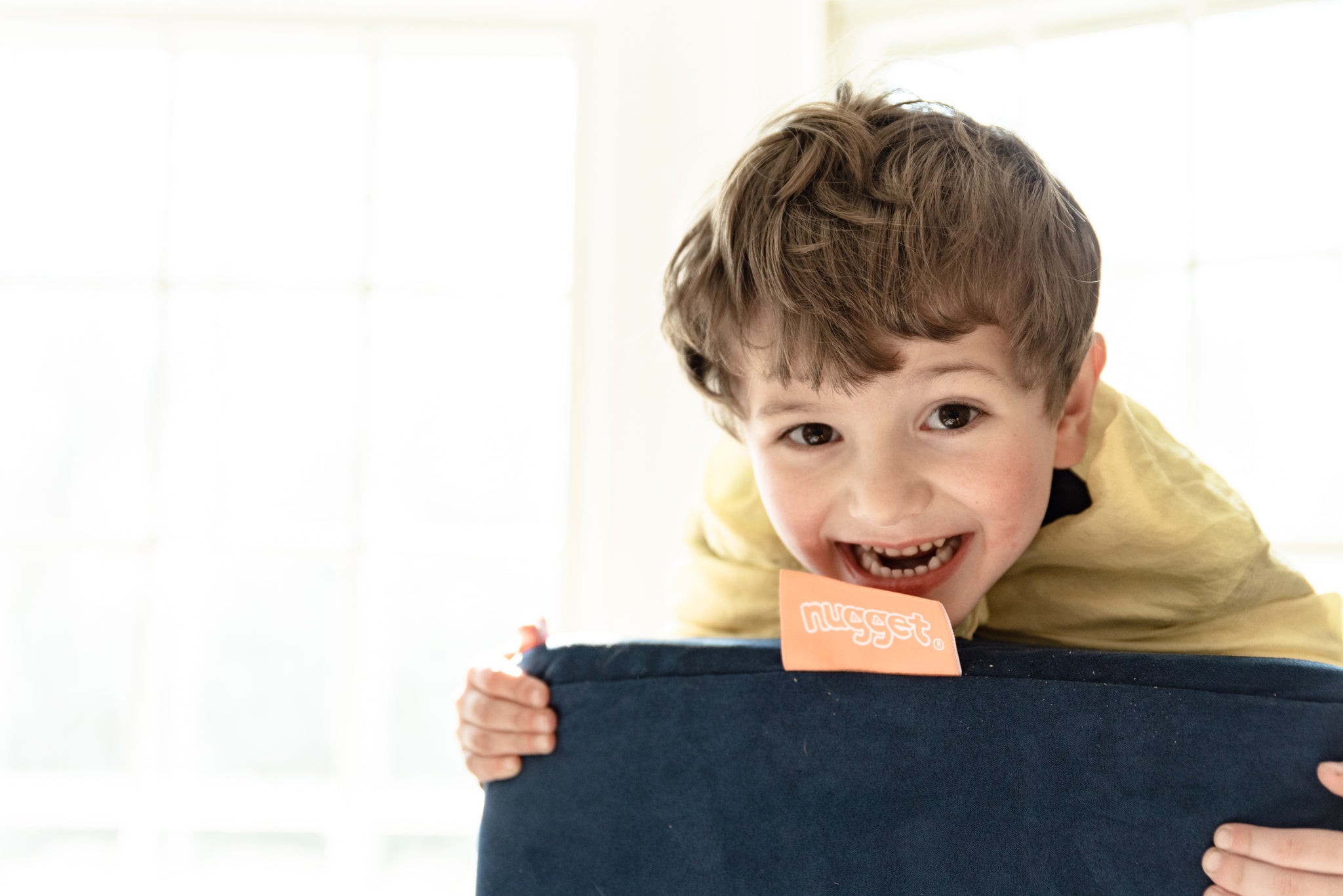 Child leans forward on a Nugget pillow, grinning at camera