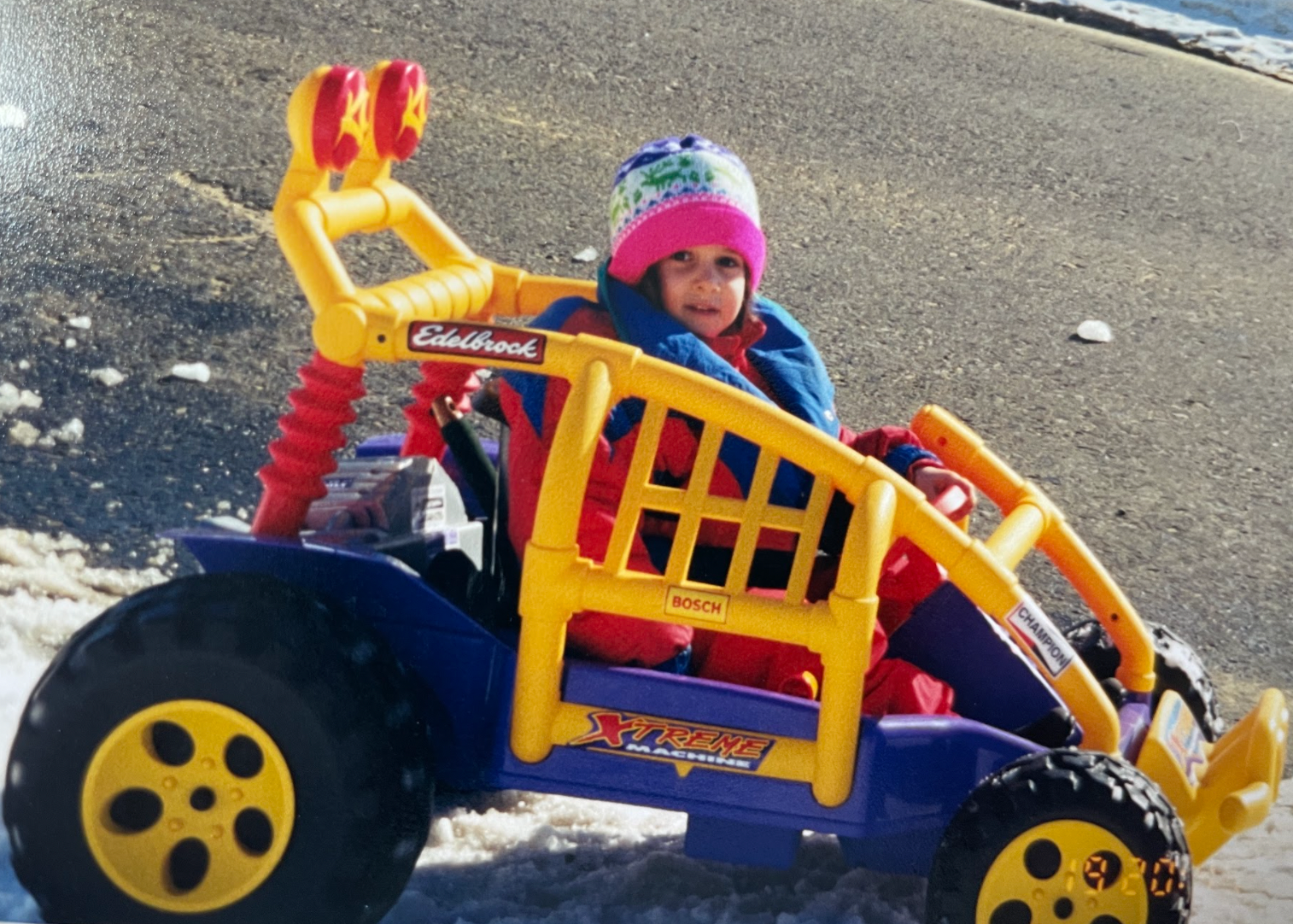 Young Ghenwa bundled up in a hat and big jacket, driving a toy car on a snowy road