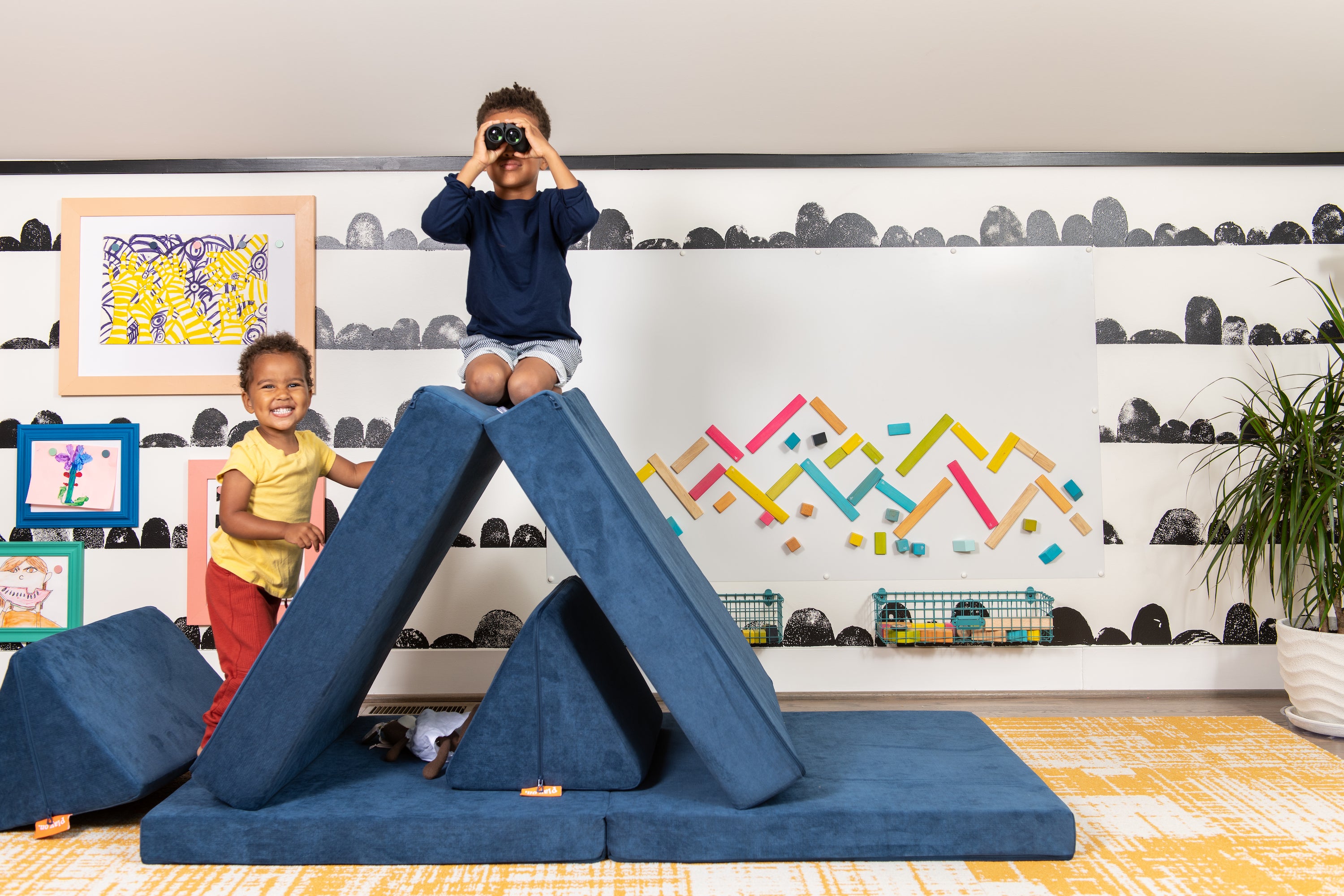 Two kids playing on a Nugget in a grOH! designed playroom. A colorful wallpaper and toys are aligned on the backwall, with a small plant in the bottom righthand corner.