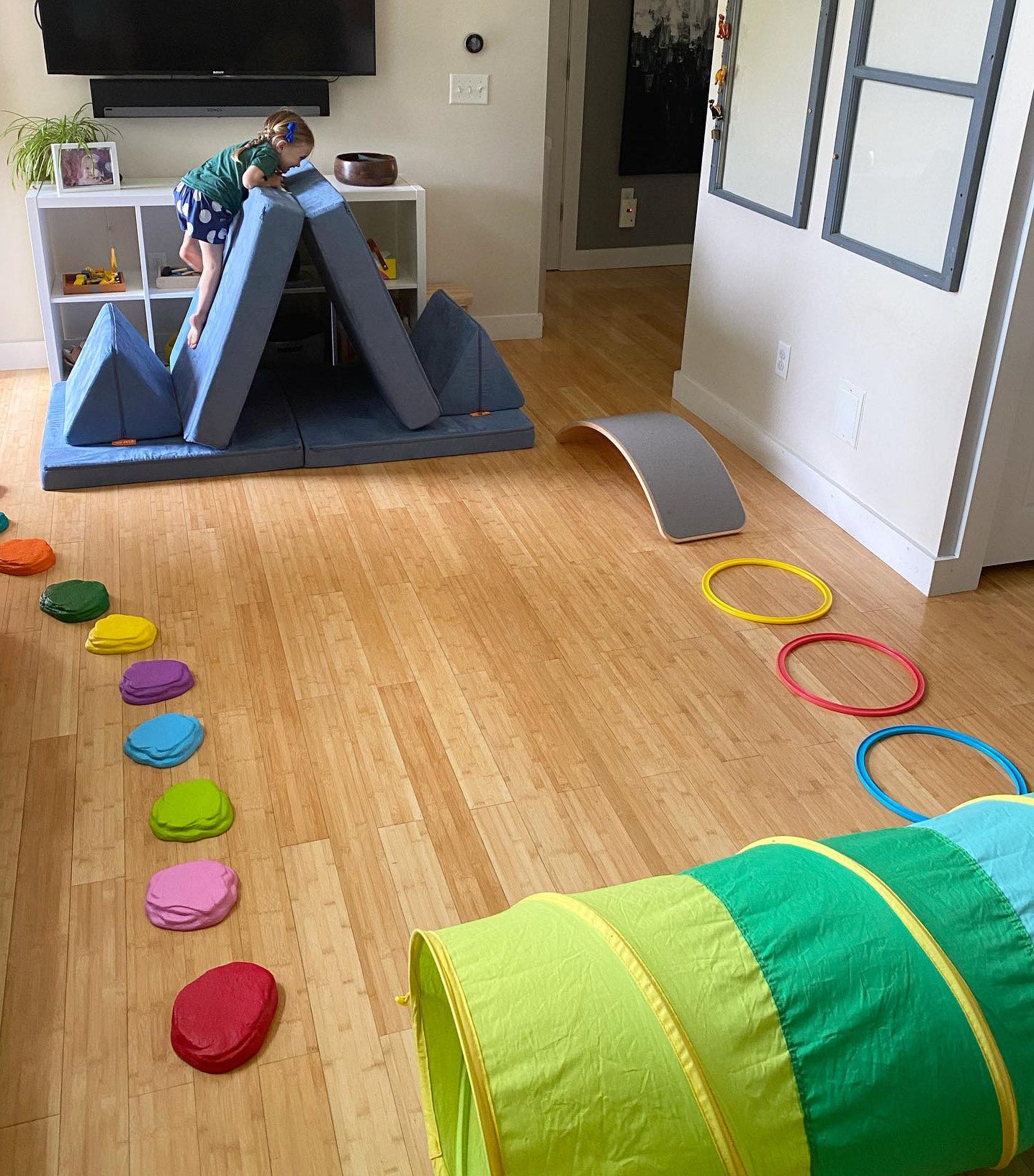 Child climbing up a Nugget base folded in two like a mountain peak, with a tunnel, stepping stones, and hula hoops set up around as an obstacle course