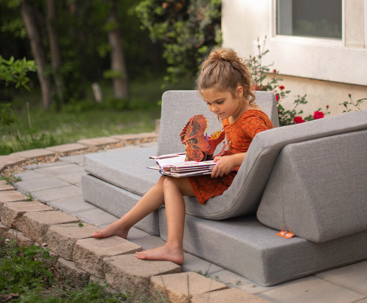 A child sits on the Outdoor Nugget on a patio walkway, reading a pop-out dinosaur book