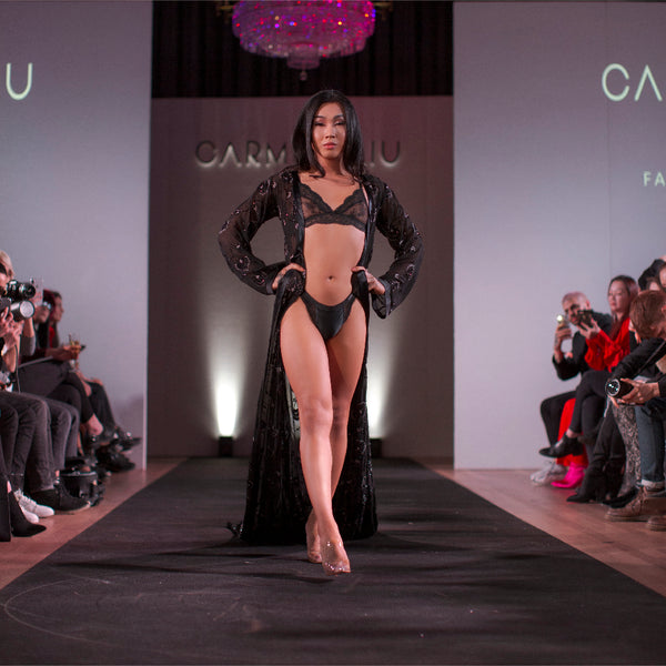 A model in black lingerie and a robe stares forward as they walk down a runway with spectators and photographers on either side.