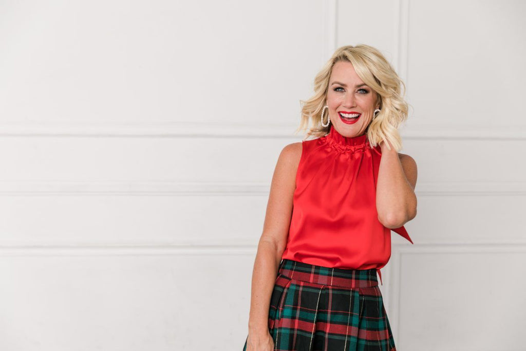 Woman wearing red blouse and plaid skirt, with grey wall in the background.