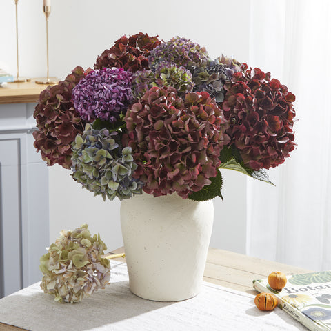 A bouquet of deep burgundy, purple and green dried hydrangeas arranged in a large vase on a table.