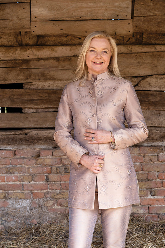 A mature blonde woman wears a pale pink suit with a nehru jacket.